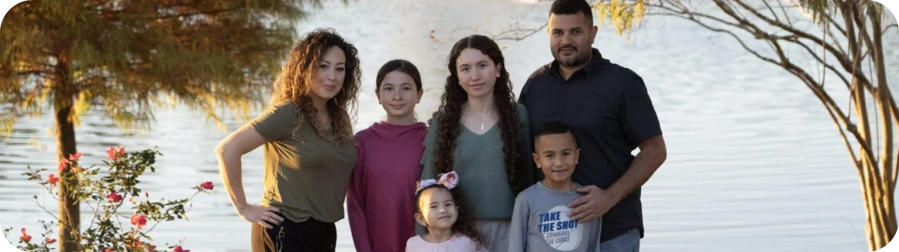 Family of six posing by a serene lake, surrounded by trees and flowers. They are smiling, conveying warmth and togetherness in a natural setting.