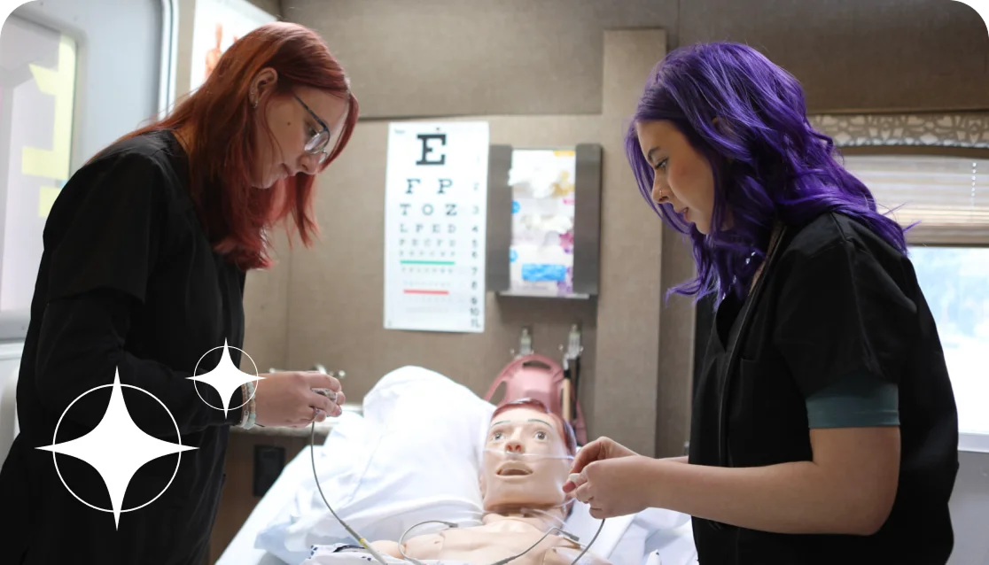 Two women with red and purple hair, wearing black scrubs, practice medical procedures on a mannequin in a clinical setting. An eye chart is visible.