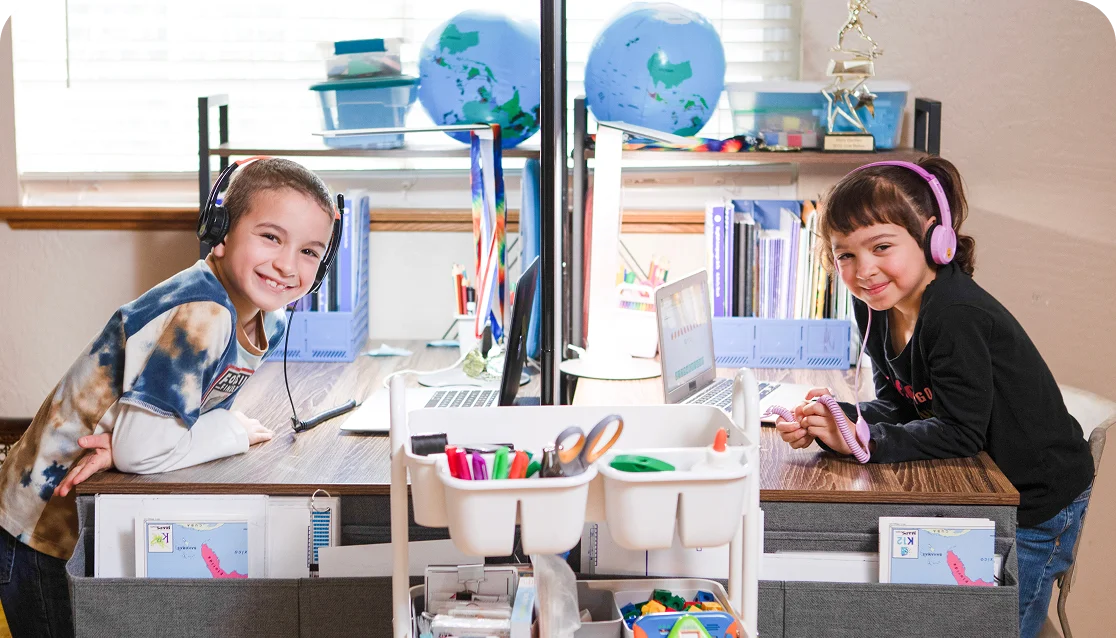 Two children wearing headphones smile while sitting at a study desk with laptops. The desk is organized with school supplies, books, and a globe in the background, conveying a cheerful learning environment.