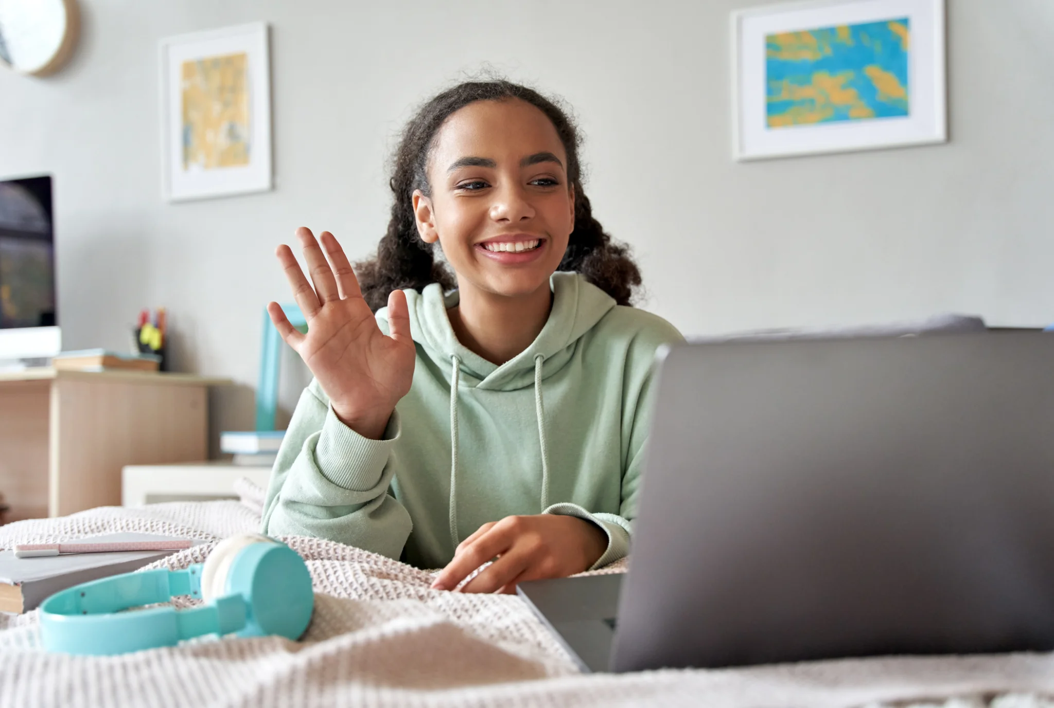 Una mujer sonriente con una sudadera con capucha verde saluda a su computadora portátil en una habitación acogedora. Los auriculares, los libros y el arte colorido crean un ambiente acogedor y optimista.