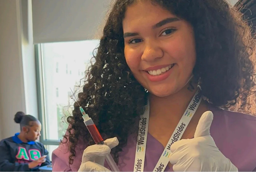 Mujer joven sonriendo con confianza, usando guantes y sosteniendo una jeringa llena de líquido rojo. Ella está en una habitación luminosa, otra persona es visible en el fondo.