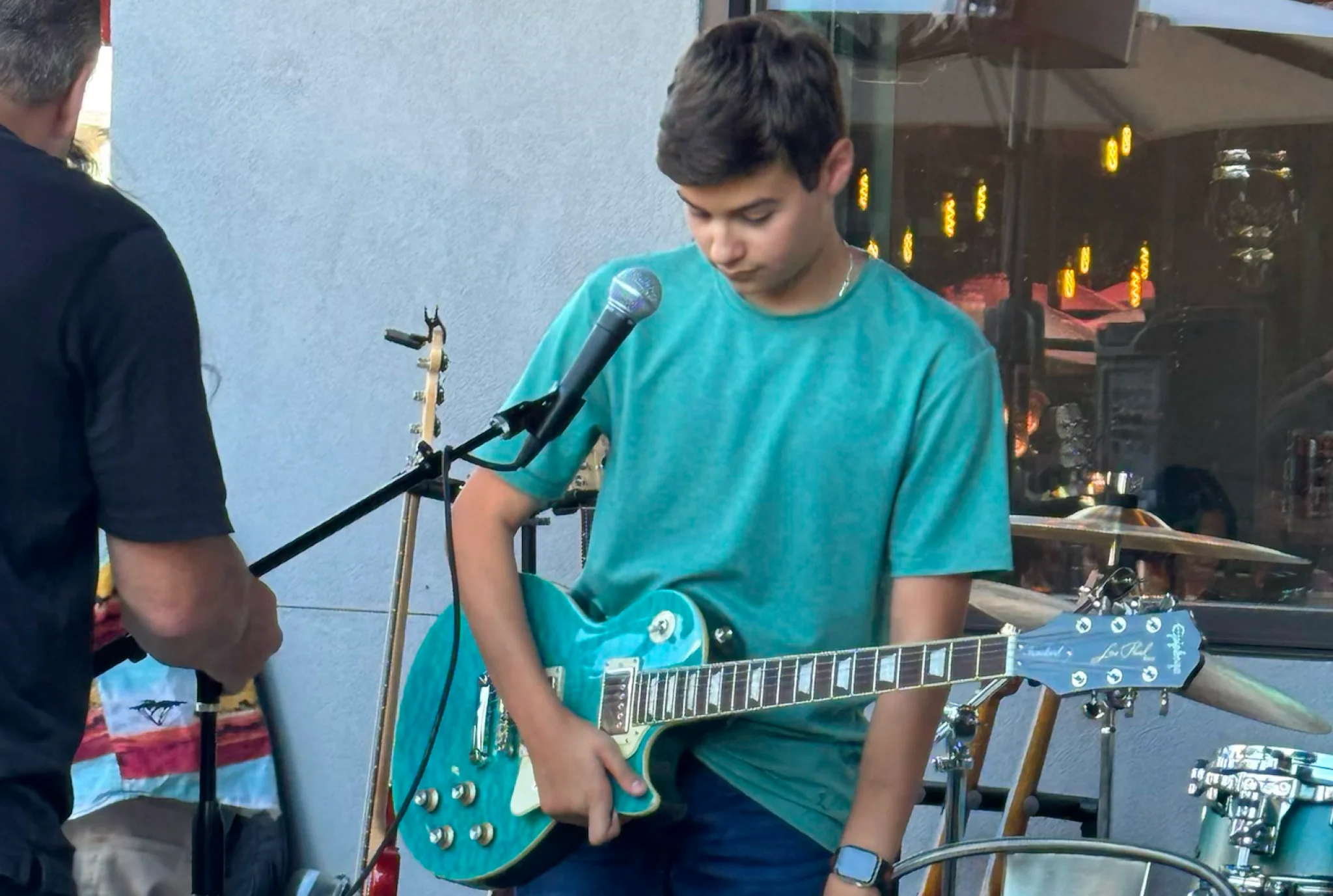 Young boy in a teal shirt tuning a blue electric guitar on stage. A microphone and drum set are nearby. Casual, focused atmosphere.