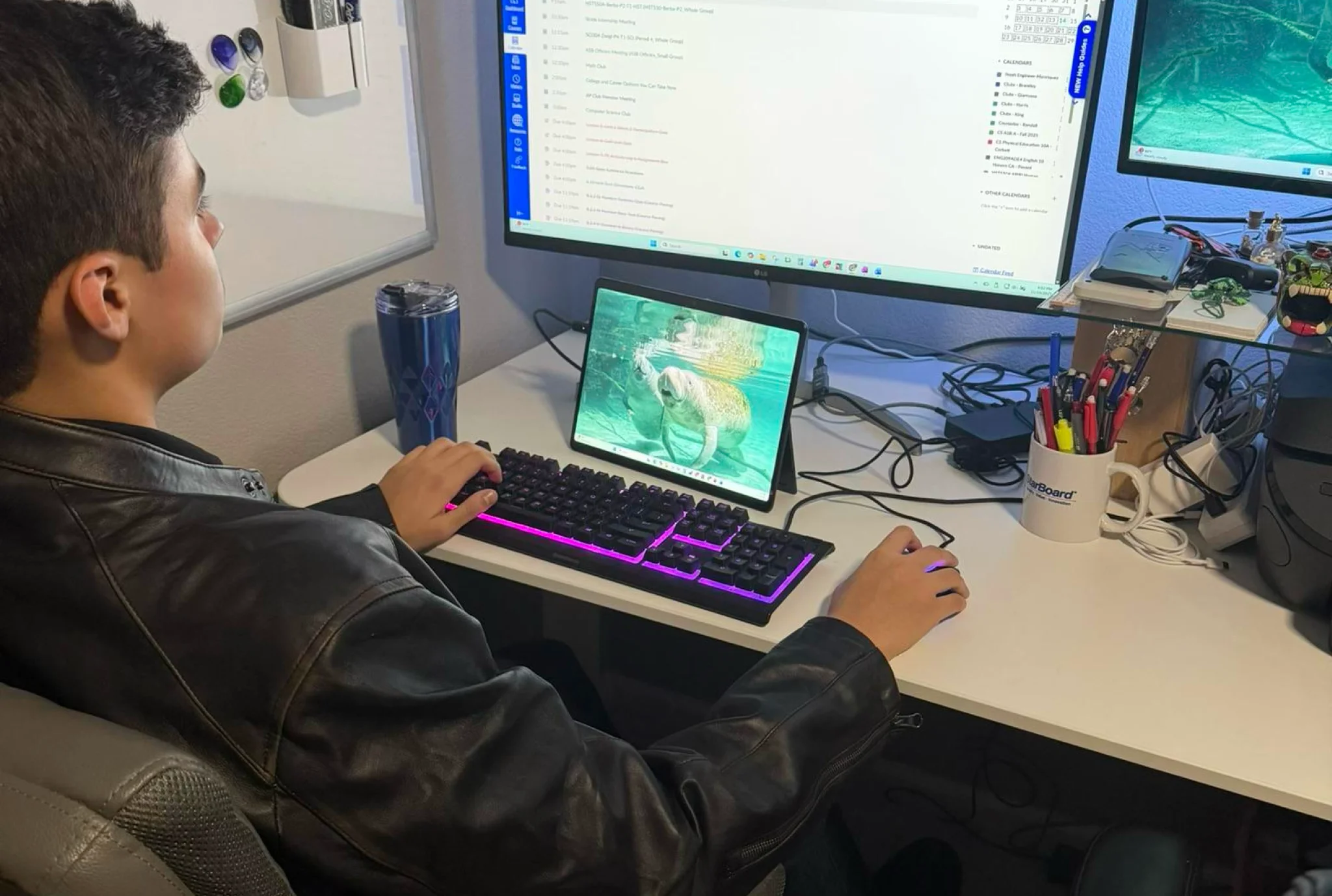 A person wearing a leather jacket sits at a desk, using a computer with a dual-monitor setup and a tablet displaying a fish. The scene is focused and productive.