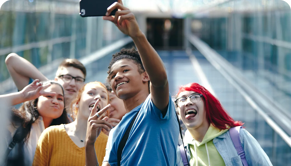 A diverse group of six young people smile and pose for a selfie on an escalator. The atmosphere is joyful and lively, capturing a moment of friendship and happiness.