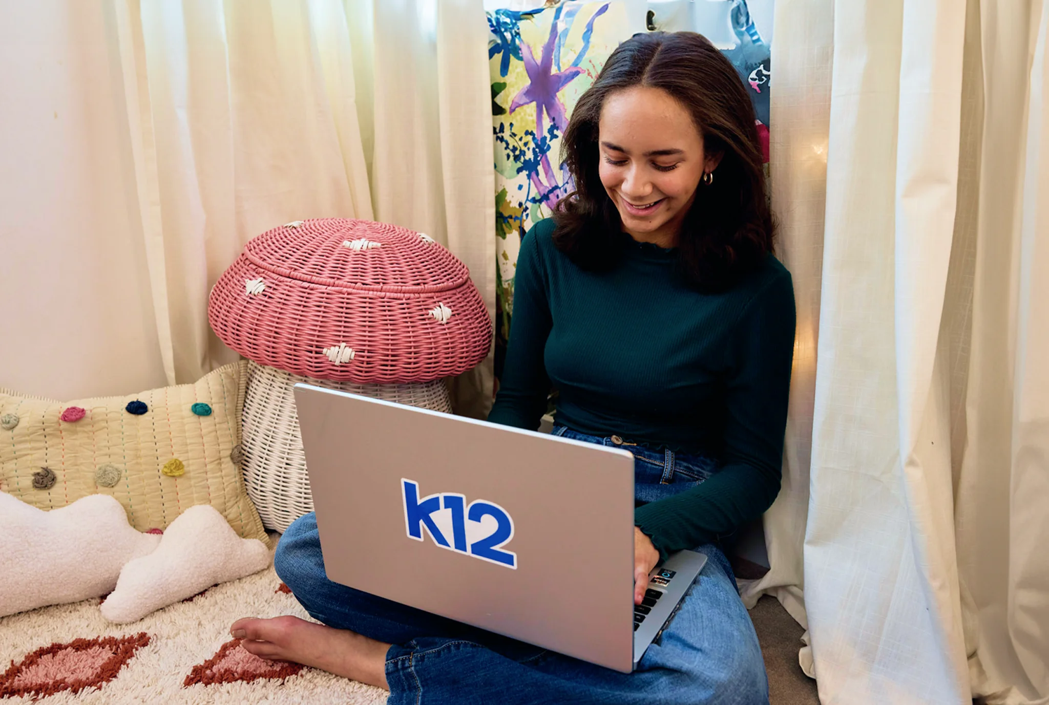 Una mujer joven se sienta con las piernas cruzadas, sonriendo a una computadora portátil con "k12" en ella. Ella está en un espacio acogedor y decorado con un cojín rosa en forma de hongo.