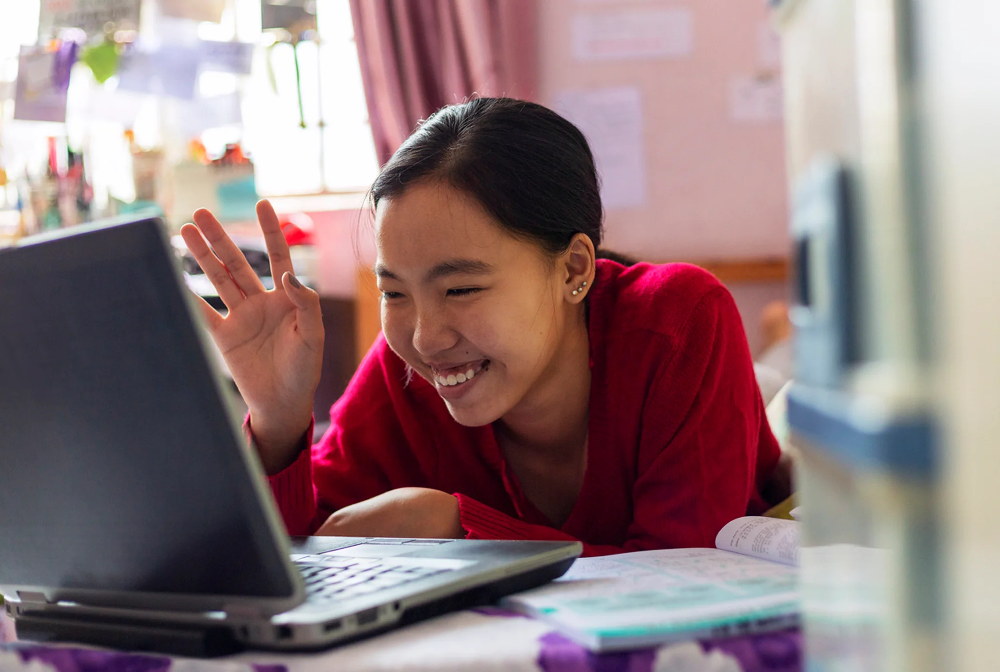 A young person in a red sweater smiles and waves at a laptop screen, creating a friendly and engaged atmosphere in a cozy, decorated room.