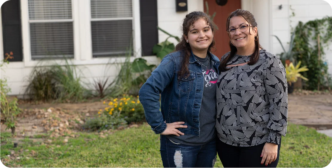 A woman and a teenage girl stand smiling in front of a house with a garden. The tone is warm and familial, with a sense of togetherness.