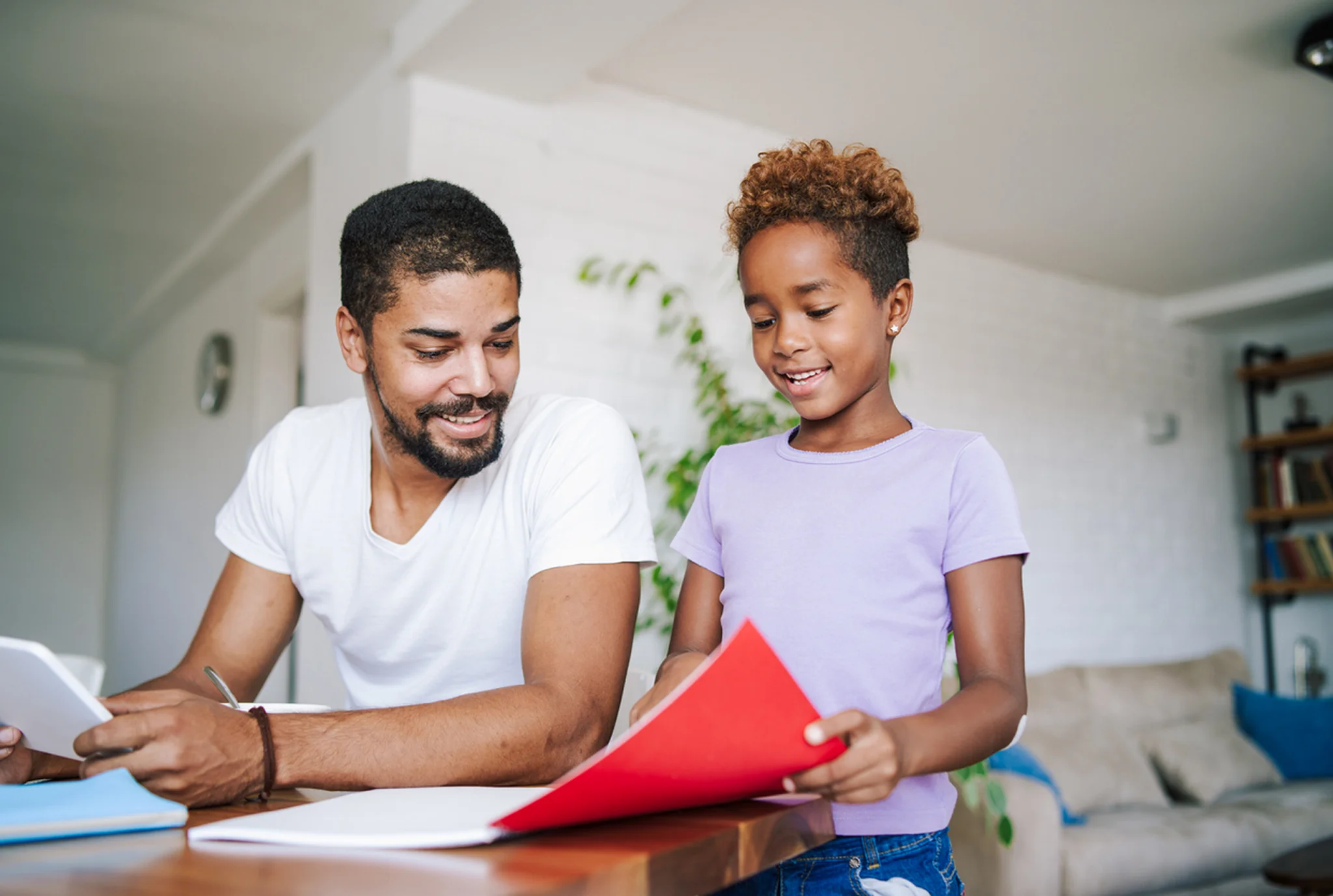 A man and a child at a table, smiling. The man holds papers, while the child in a purple shirt shows a red folder. The atmosphere is warm and joyful.