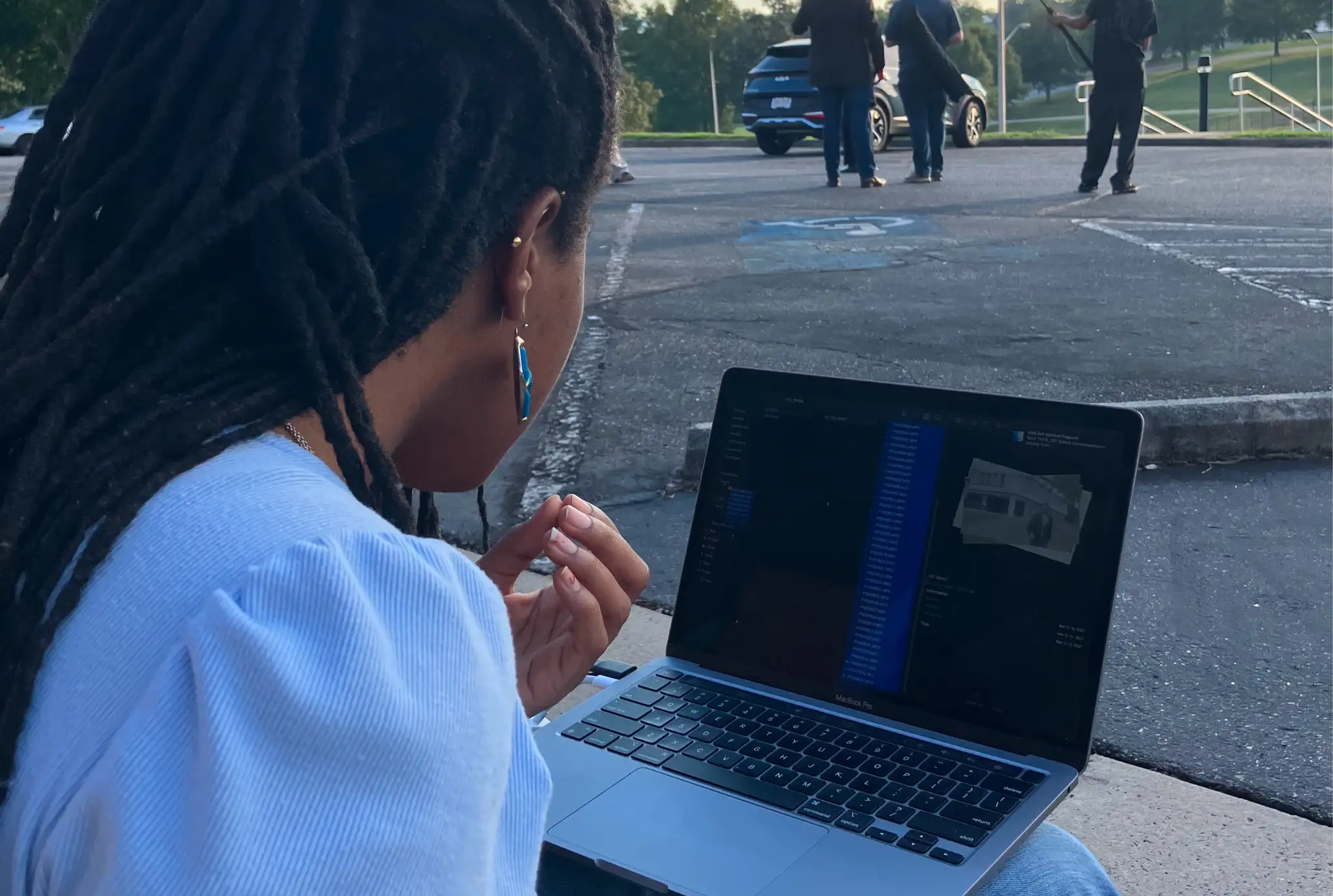 A person with long hair and blue earrings sits outdoors using a laptop showing a 3D image. In the background, people stand near a parked car on a road.