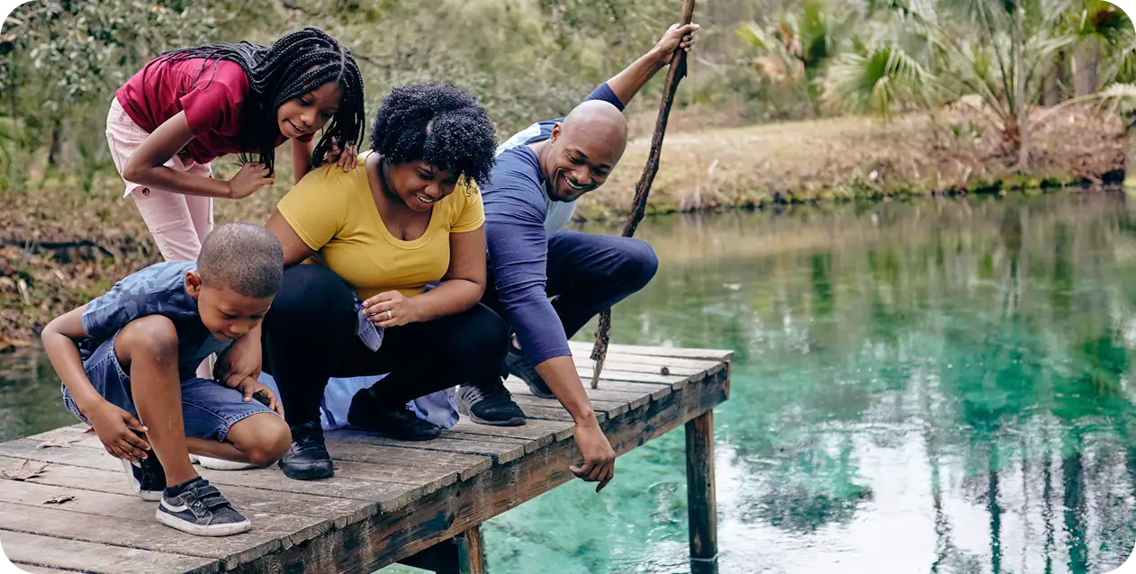 A joyful family of four, on a wooden dock, peers into a clear pond. Lush greenery surrounds them, creating a serene and playful atmosphere.