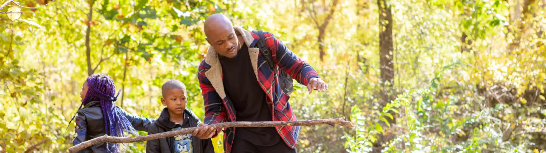 A family explores a sunlit forest in autumn. A man in a plaid jacket interacts with a large stick, with two children observing. Scene is serene and warm.