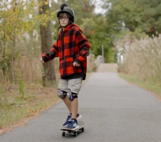A young boy wearing a red plaid jacket and shorts skateboards down a tree-lined path, wearing a helmet and knee pads for safety. The scene is set in an autumn setting, conveying a sense of adventure and focus.