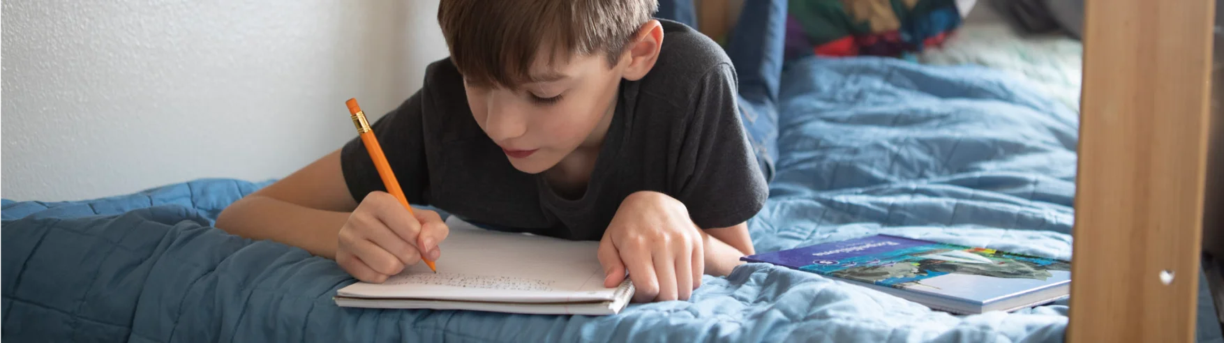 Young boy lies on a blue bedspread, focused on writing in a notebook with a pencil. A textbook lies beside him, suggesting study time.