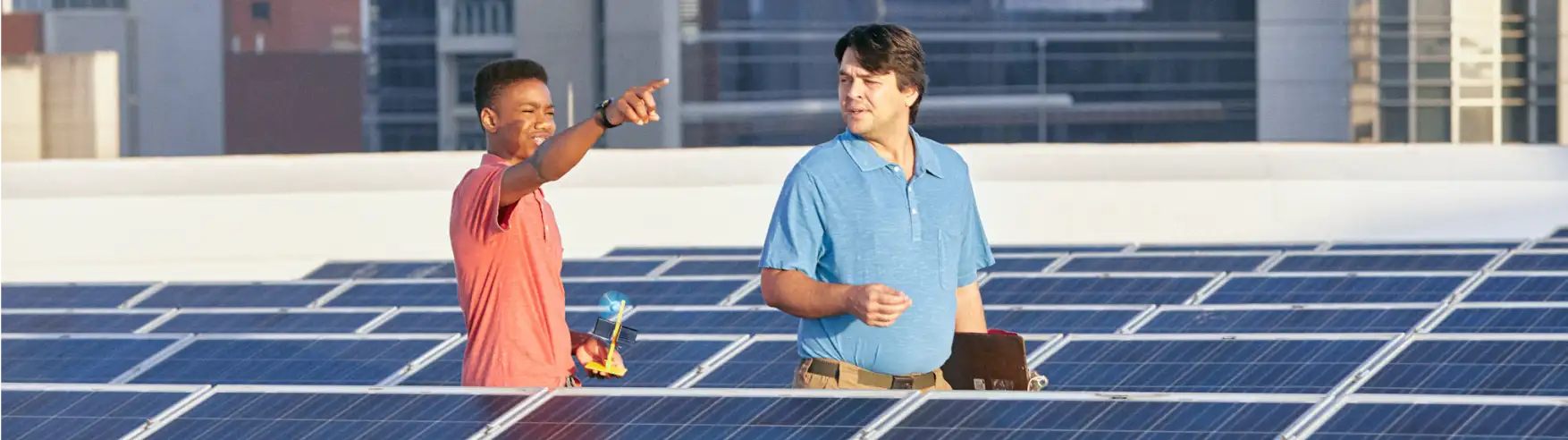 Two people stand on a rooftop covered with solar panels. One points ahead enthusiastically, while the other holds a clipboard, both smiling.