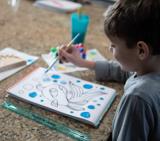 A young boy is joyfully painting a cartoon fish in a notebook, surrounded by art supplies on a countertop. A blue cup and paints are nearby.