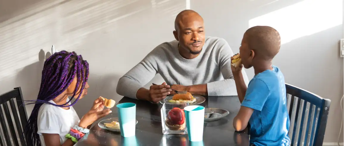 A man and two children share a meal at a table. There are plates with food, a clear vase with apples, and blue cups. The mood is warm and familial.