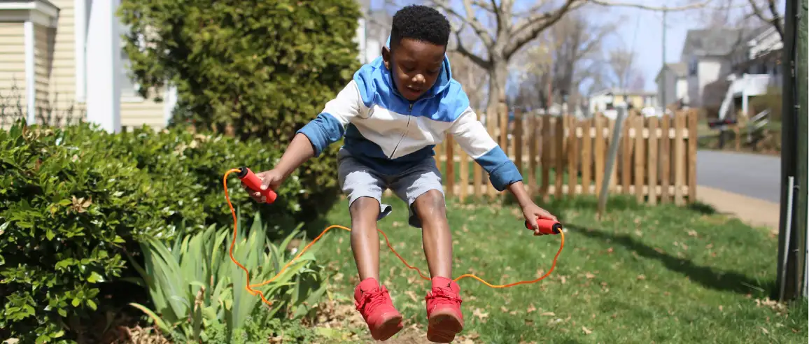 Un niño salta con una cuerda roja para saltar en un patio iluminado por el sol, vistiendo una sudadera con capucha azul y blanca y zapatos rojos, transmitiendo alegría y energía en un día brillante.