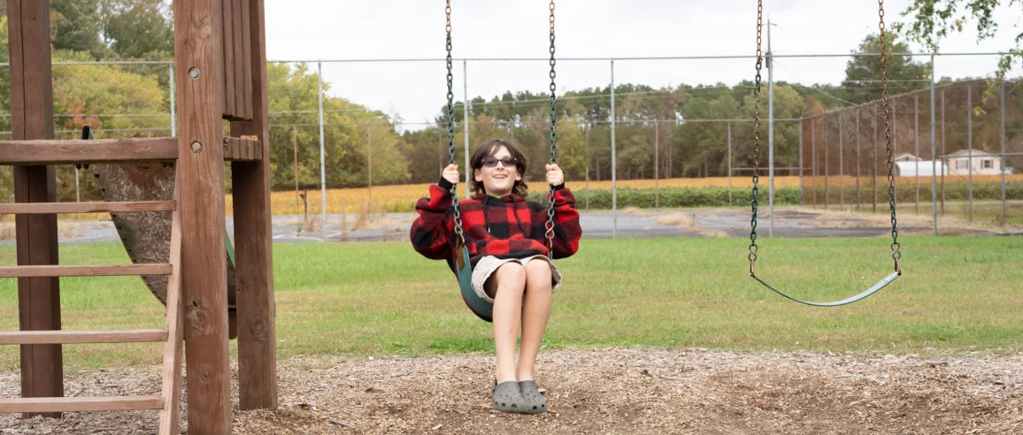 A child in sunglasses joyfully swings on a playground set, wearing a red and black plaid jacket. The background features trees and a grassy field.