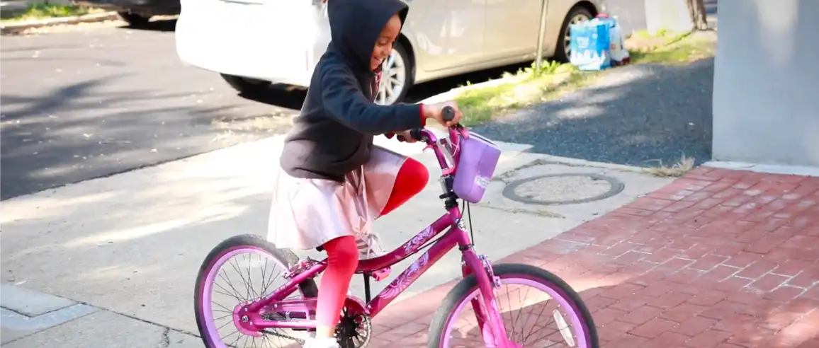 A young child in a black hoodie and pink skirt rides a bright pink bicycle on a sunny sidewalk. The scene is cheerful, evoking a sense of joy and playfulness.