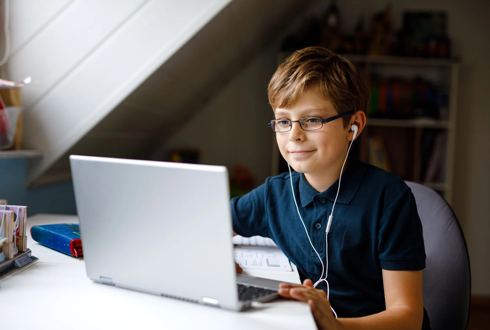 A young boy with glasses is smiling while using a laptop with earphones in a cozy, well-lit room. The scene conveys focus and a cheerful mood.