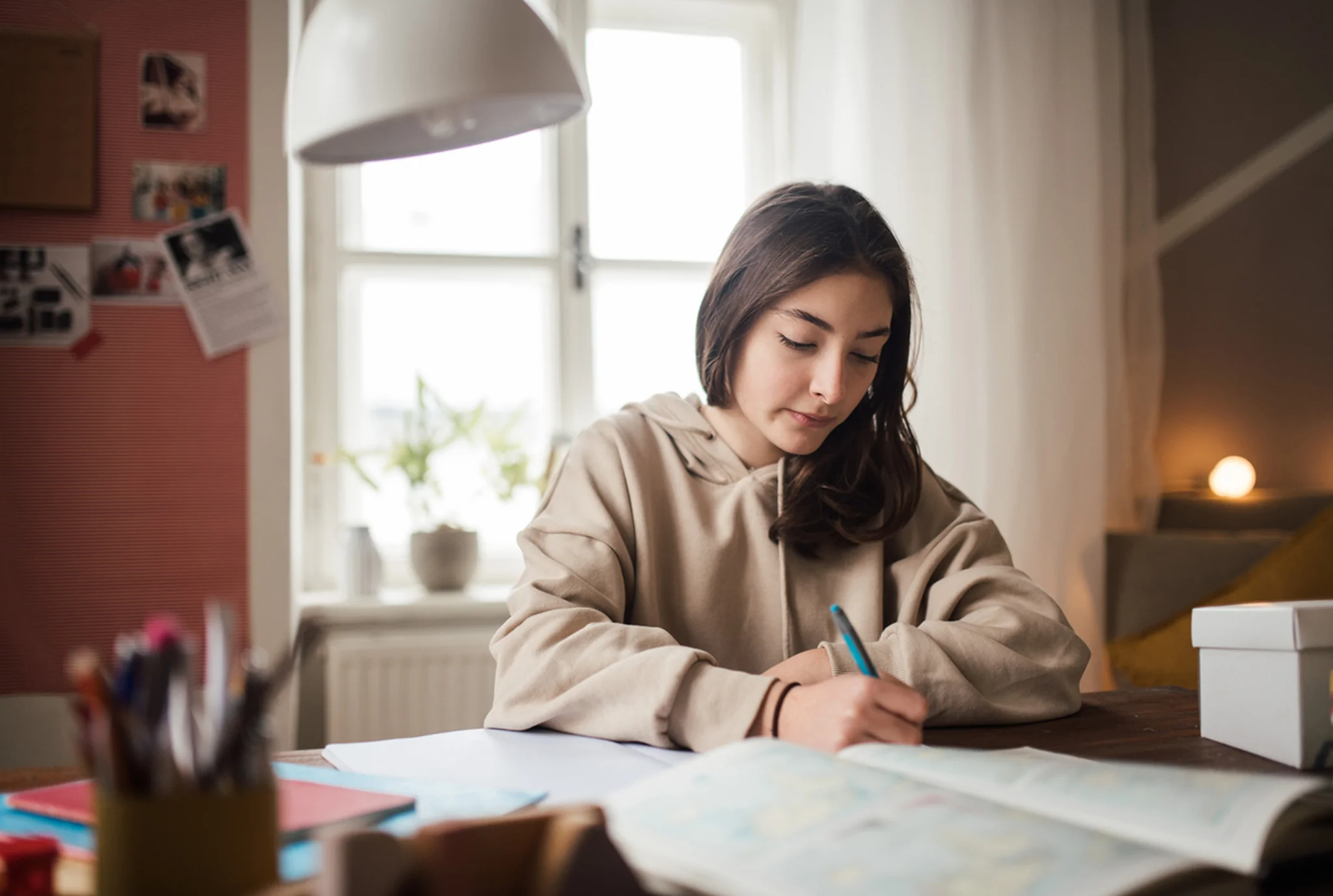 Young woman in a beige hoodie working at a desk with papers and pens, focused and calm. A bright window and cozy room create a peaceful vibe.
