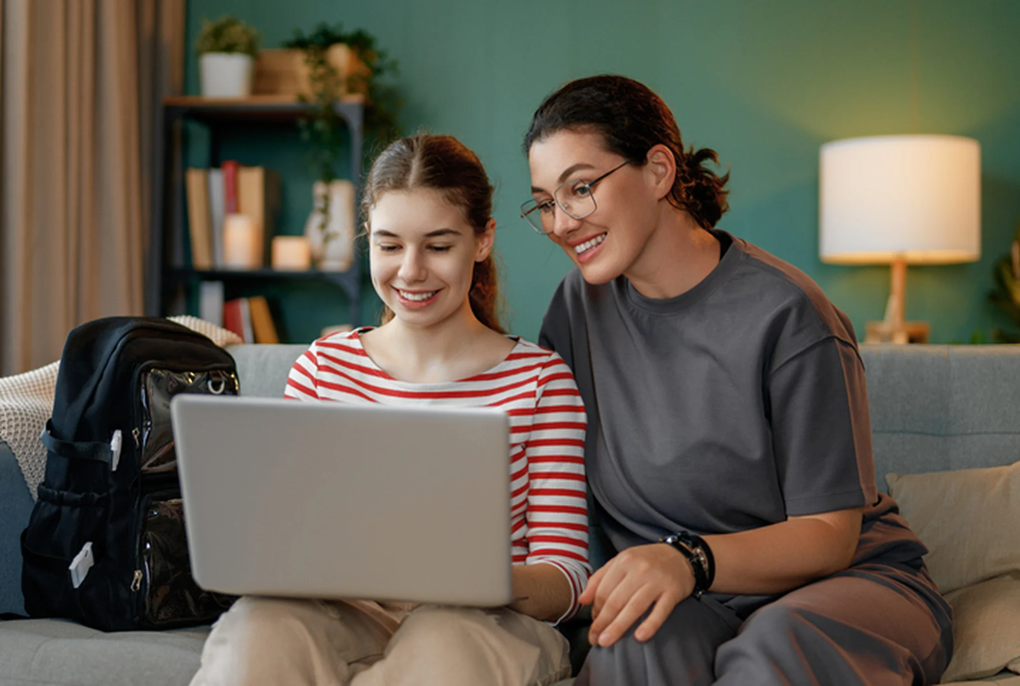 A mother and daughter sit on a cozy sofa, smiling while looking at a laptop. The room is warmly lit, with a lamp, books, and a backpack nearby.