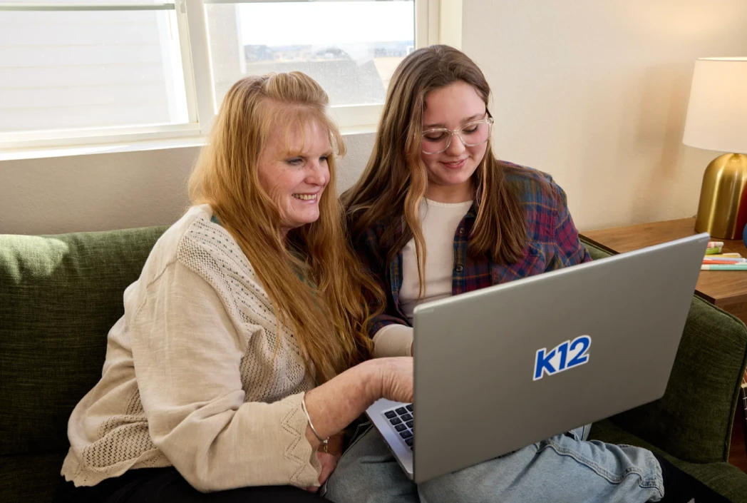 Two people sit on a green sofa, smiling and engaging with a laptop displaying "k12." A lamp and window are in the background, evoking a cozy, learning-focused atmosphere.