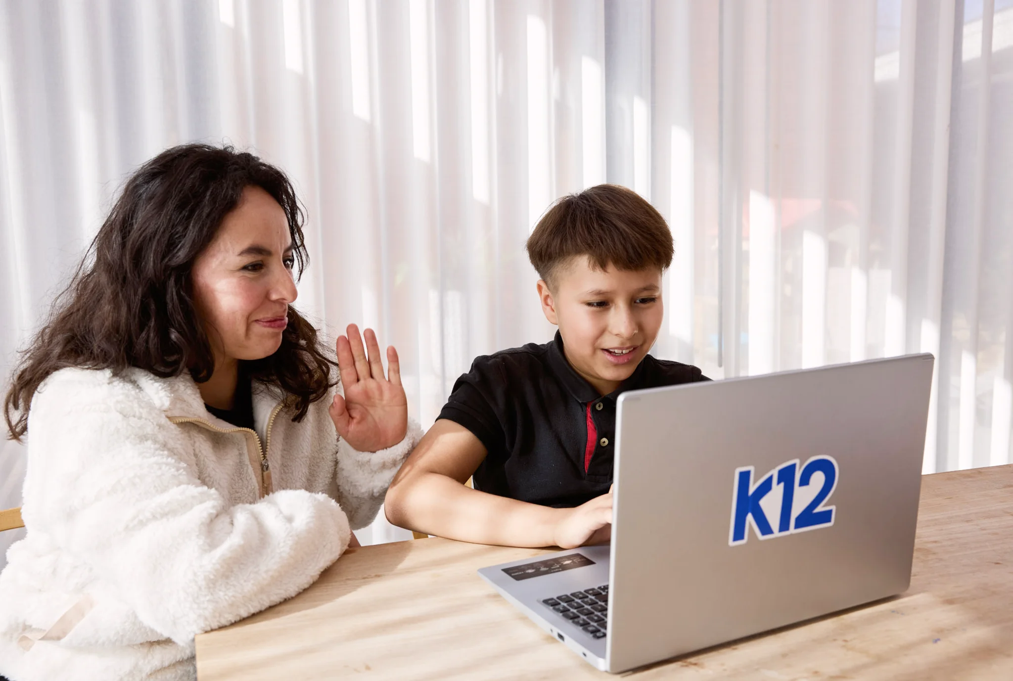 A smiling child and an adult sit at a table with a laptop displaying "k12." The adult waves warmly, suggesting a friendly, educational scene.