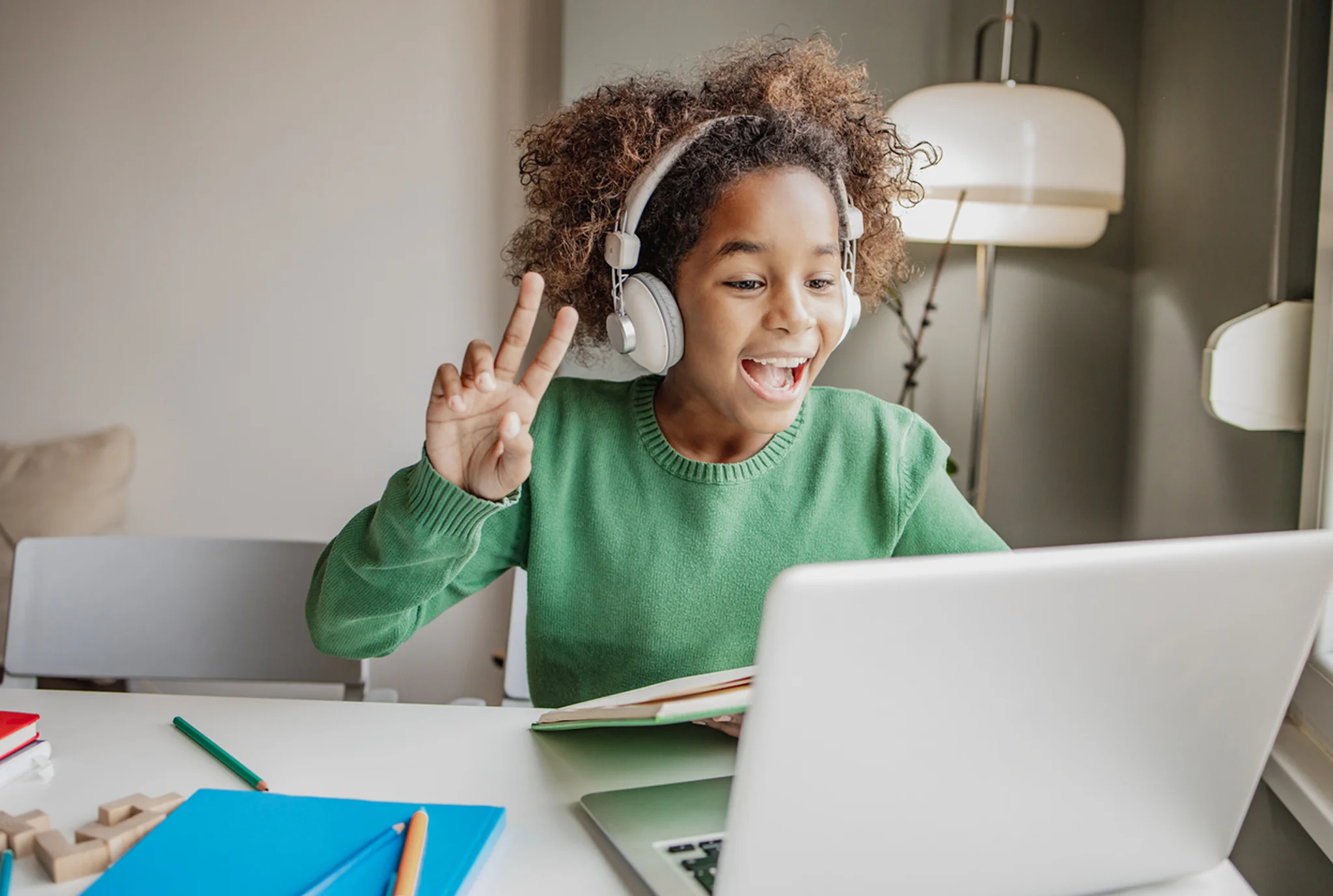 Un niño alegre con auriculares y un suéter verde saluda a una computadora portátil durante una clase en línea. La habitación es luminosa y organizada, transmitiendo emoción.