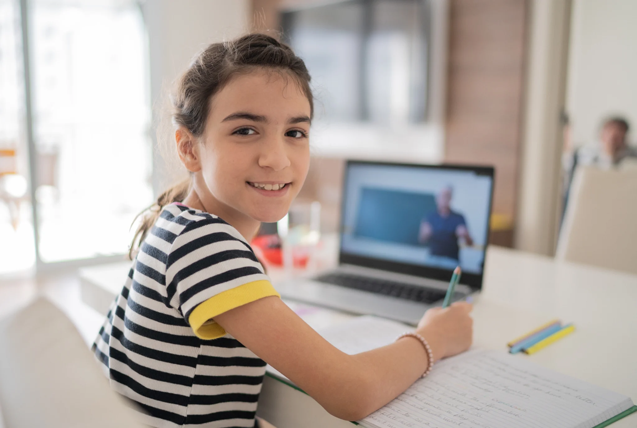 Una joven con una camisa a rayas sonríe mientras estudia en una mesa blanca. Ella está escribiendo en un cuaderno, con una pantalla de computadora portátil que muestra una lección en línea.