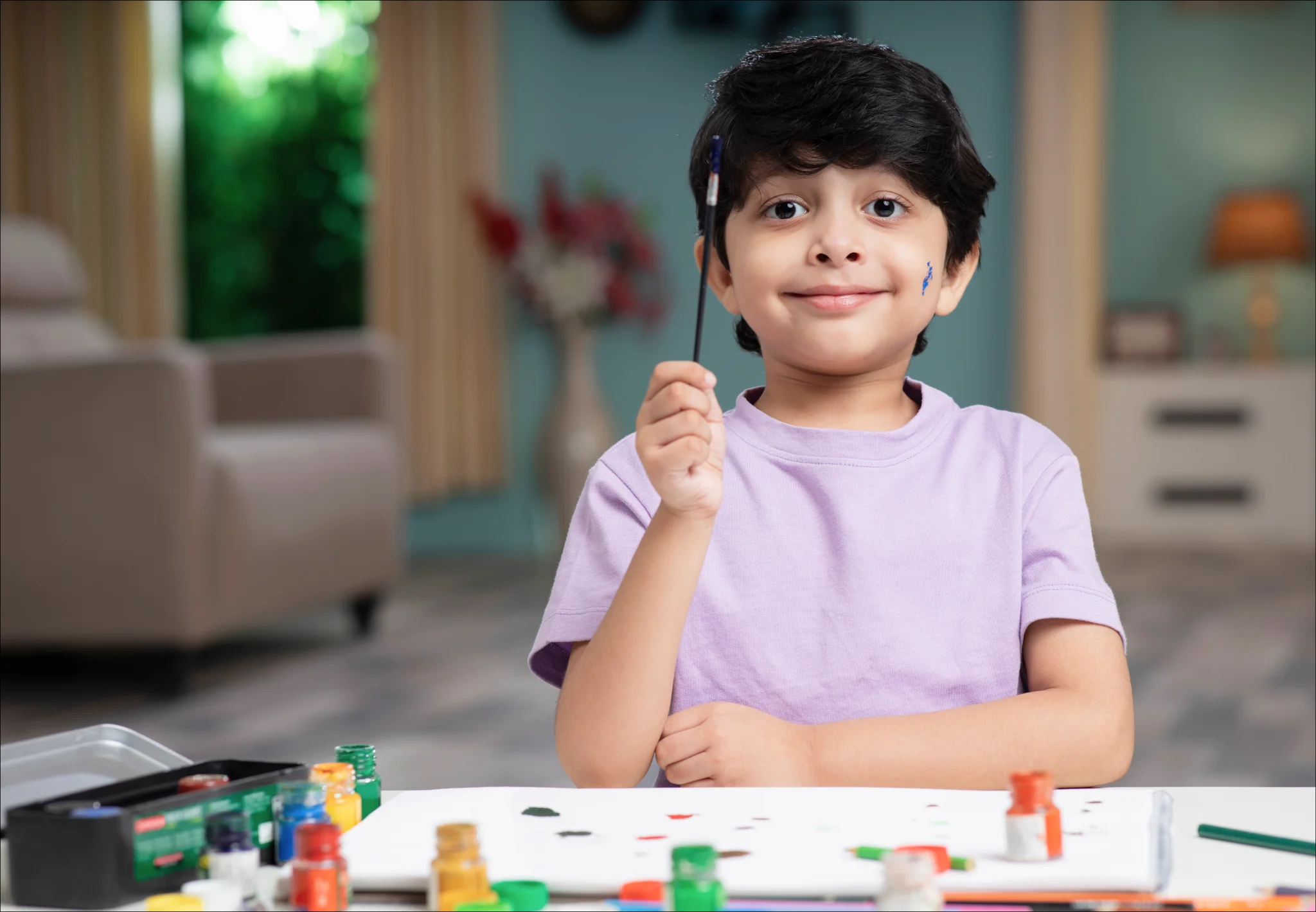 A smiling child with short dark hair in a purple shirt holds a paintbrush. They sit at a table with colorful paints, conveying creativity and joy.