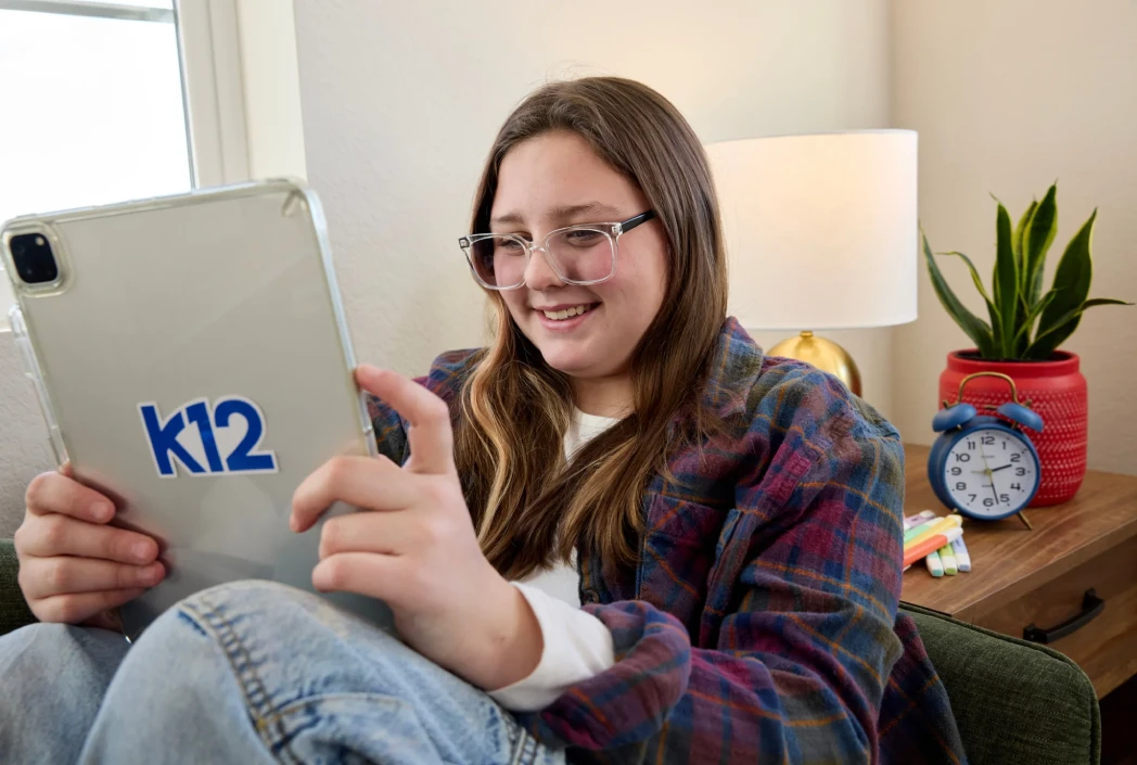 A young person with glasses smiles while using a tablet with a "k12" sticker, sitting beside a lit lamp, alarm clock, and potted plant in a cozy room.