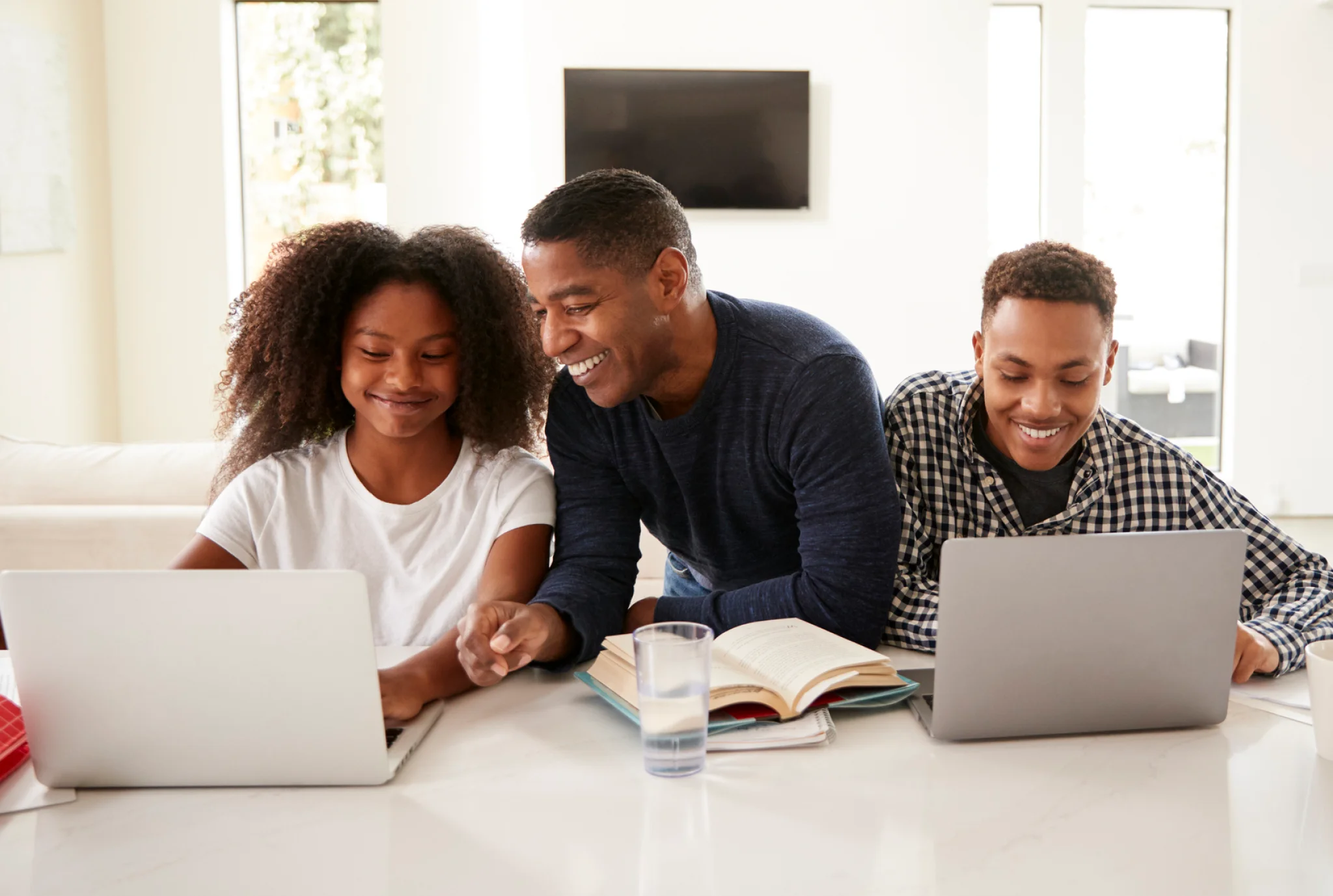 A man and two teenagers, smiling, sit at a table with laptops and an open book, suggesting a positive learning or study environment at home.
