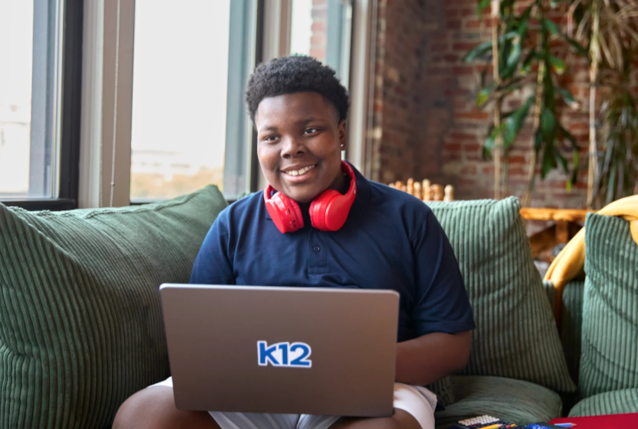 Smiling child with red headphones sits on a couch using a laptop with "k12" on it. Sunlight filters through a large window, creating a cozy ambiance.
