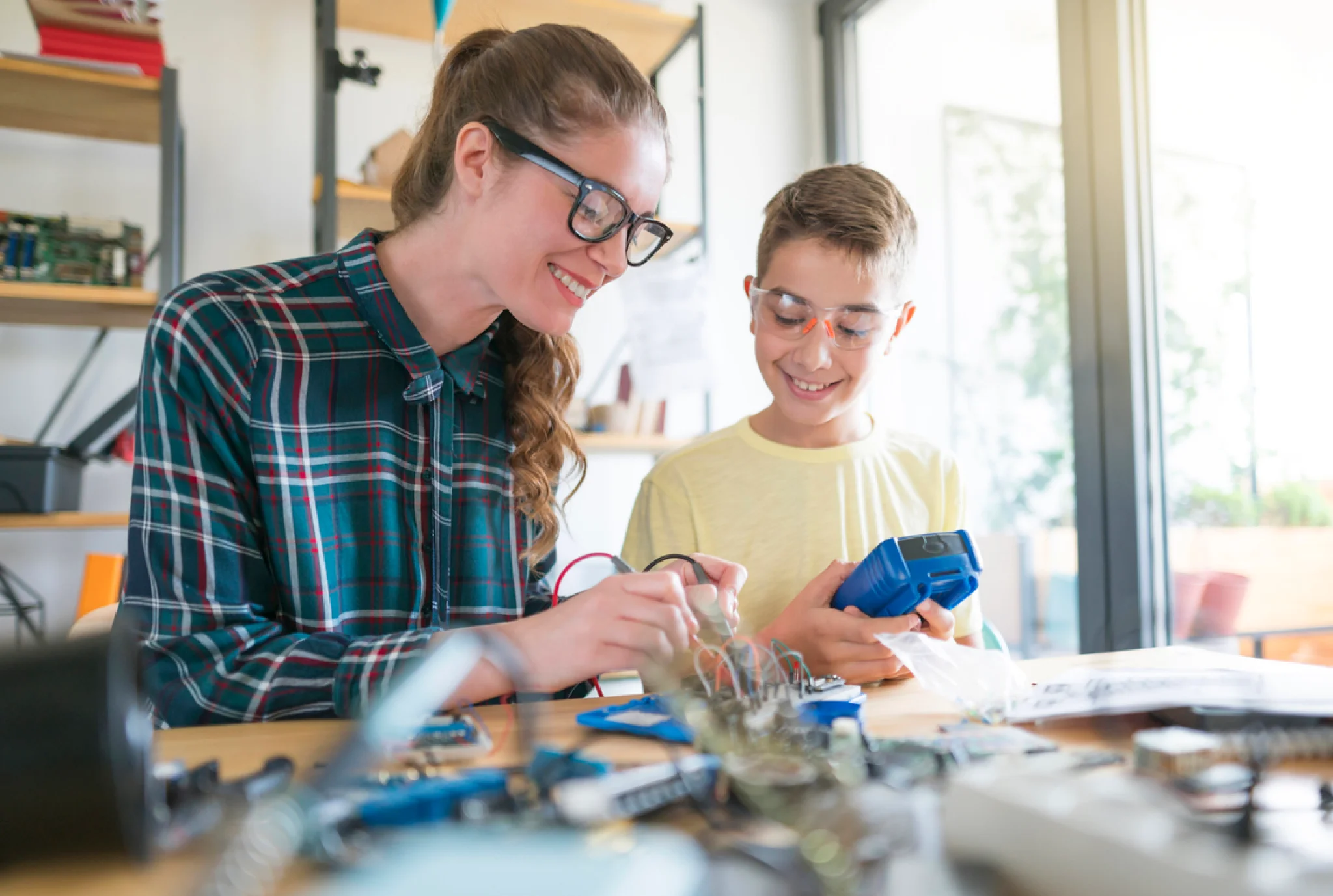 A woman and a young boy, both wearing safety glasses, engage in a fun electronics project at a sunlit table. Smiles reflect excitement and collaboration.