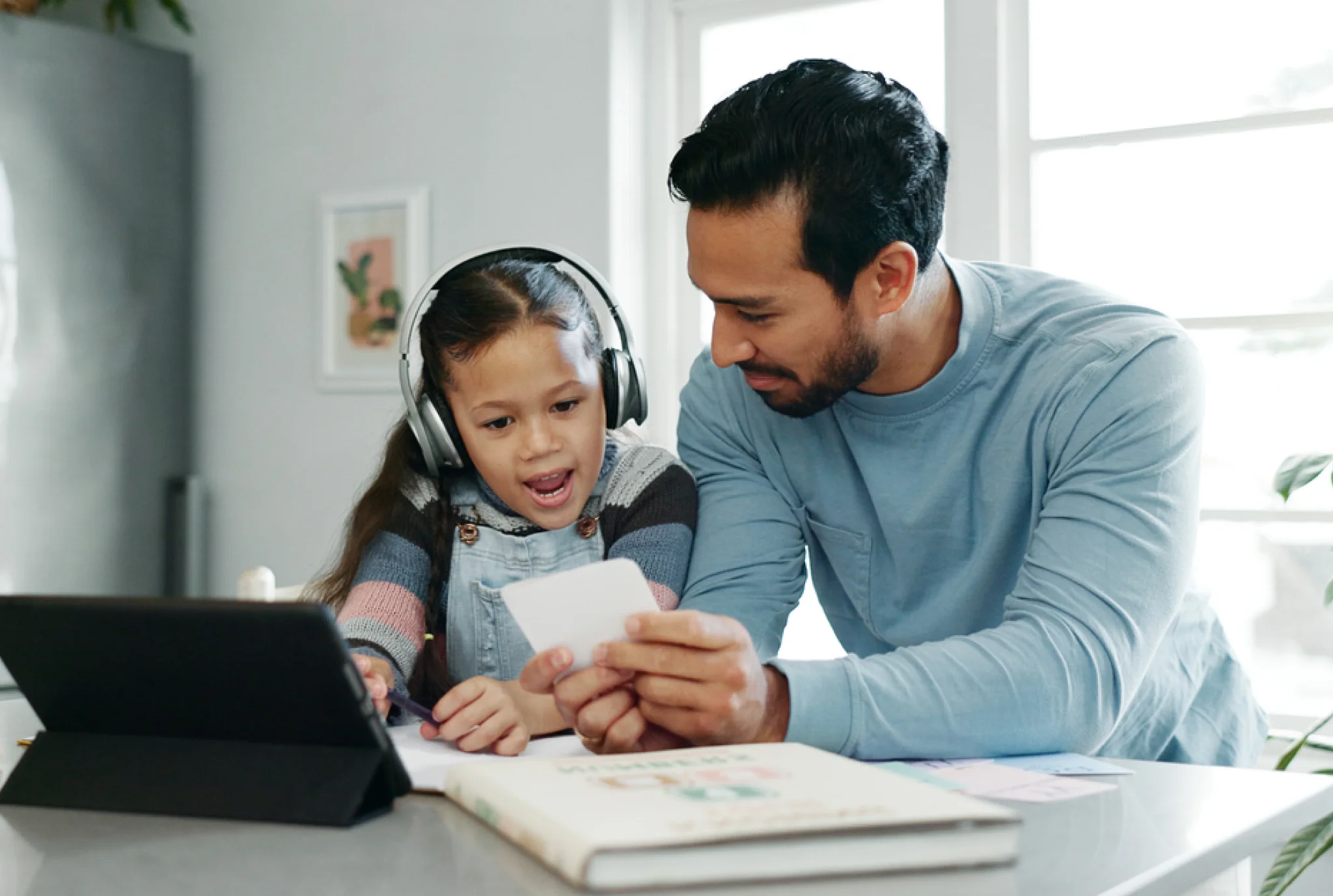 A man and a young girl in headphones sit at a table, looking at flashcards. A tablet and book are nearby. The mood is educational and focused.