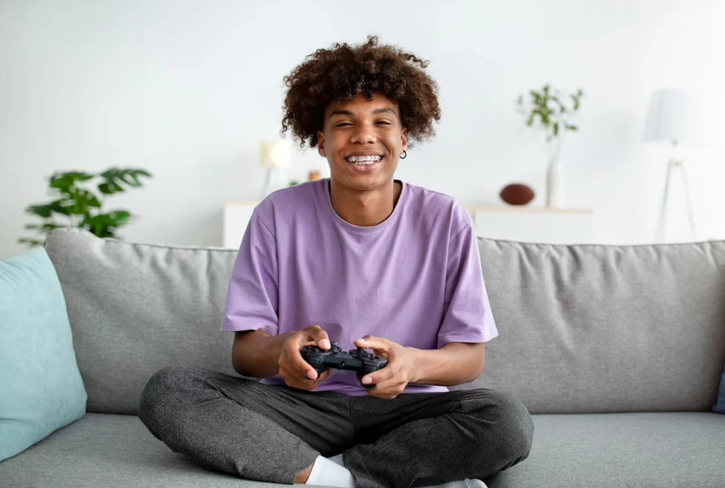 Joven con cabello rizado y una camisa morada, sonriendo mientras sostiene un controlador de juego. Se sientan con las piernas cruzadas en un sofá en una habitación luminosa y acogedora.