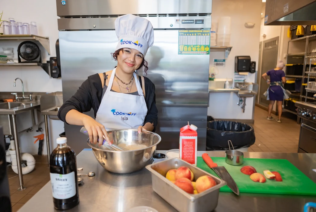 Joven sonriendo, con sombrero de chef y delantal, mezcla ingredientes en un tazón en una cocina comercial. Manzanas y utensilios de cocina están en el mostrador, transmitiendo un ambiente culinario alegre.