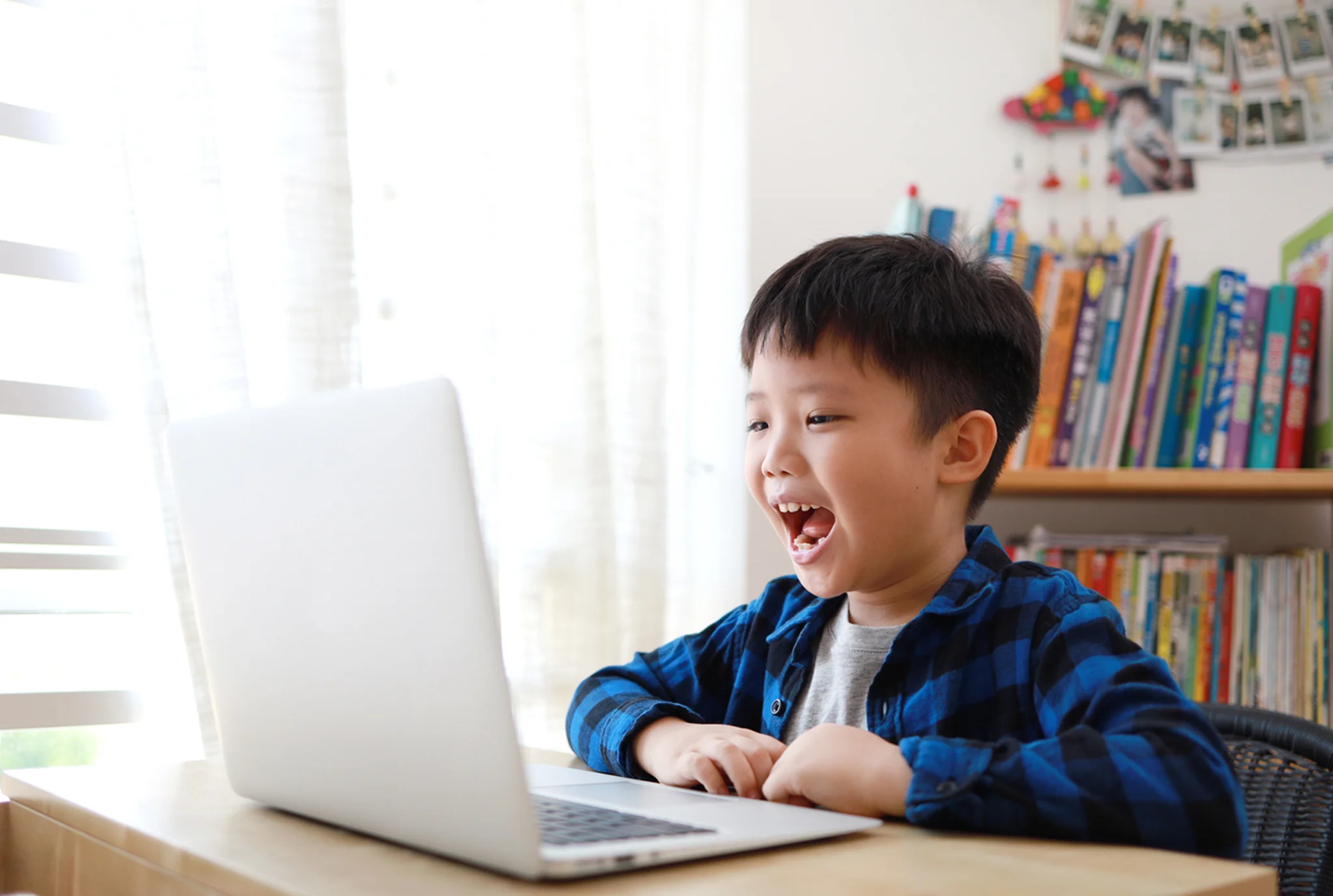 A joyful young boy in a blue plaid shirt sits at a table, happily interacting with a laptop. Sunlight filters through nearby curtains. Books and photos fill the background.