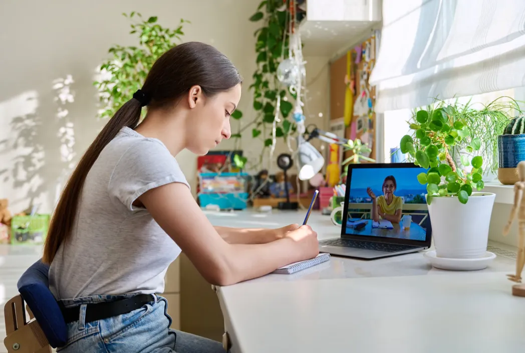 Una mujer joven se sienta en un escritorio, escribiendo en un cuaderno. Una computadora portátil con una videollamada está abierta a su lado. La habitación tiene plantas y un ambiente soleado.