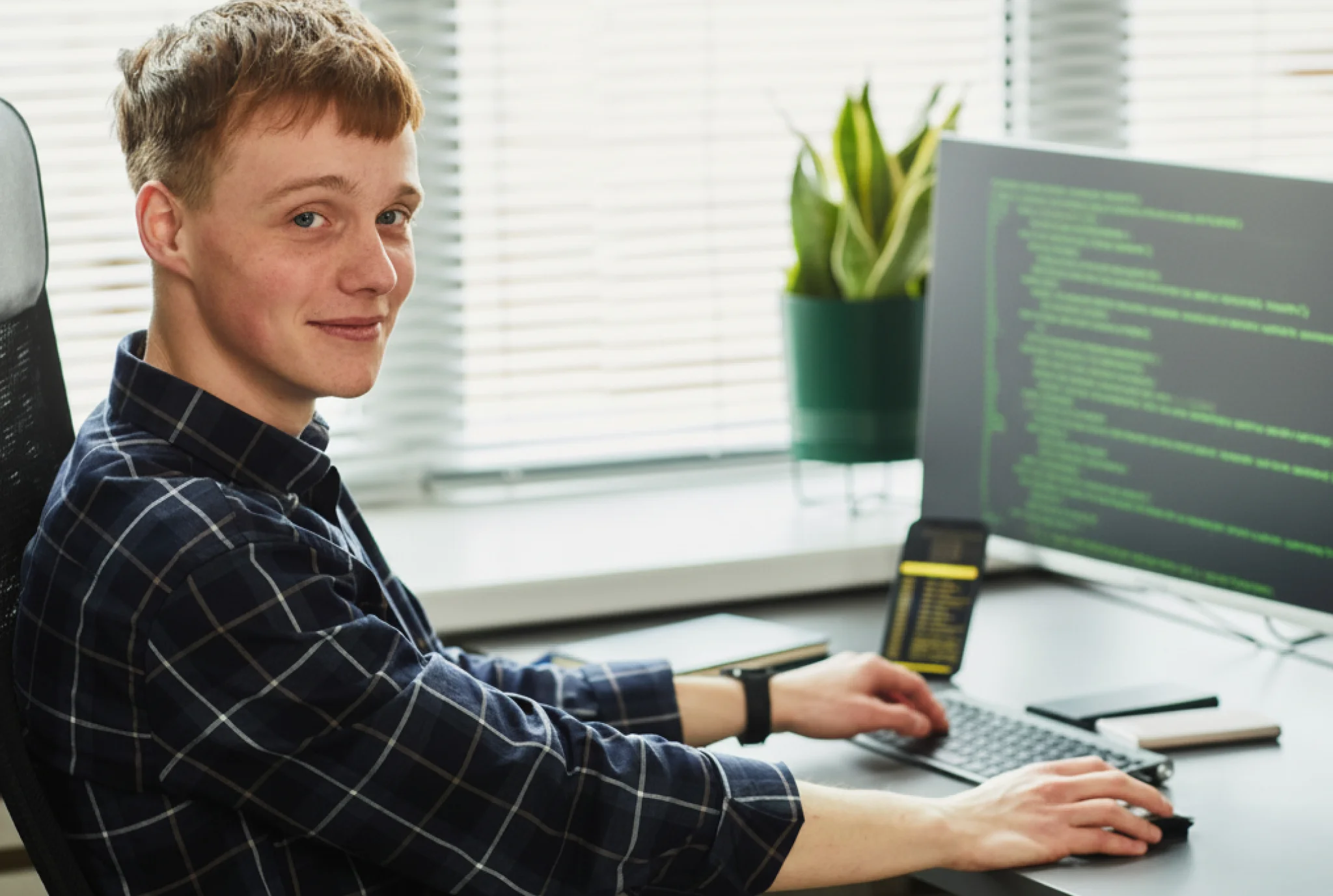 Un joven con una camisa a cuadros sonríe mientras escribe en un escritorio con un monitor que muestra el código. Una planta en maceta y persianas están en el fondo.