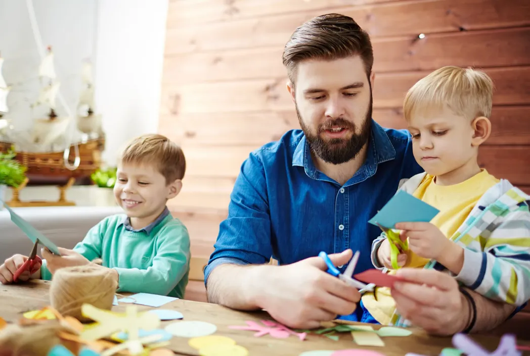Un padre y dos niños pequeños participan en una actividad de manualidades de papel en una mesa, rodeados de coloridos recortes. Todos parecen enfocados y alegres.