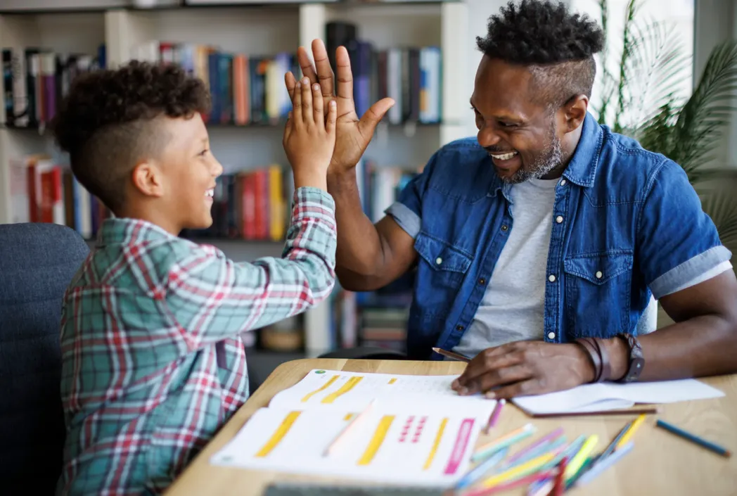 Padre e hijo sonriendo y chocando los cinco en un escritorio con papeles y lápices de colores, transmitiendo un ambiente de aprendizaje alegre y de apoyo. Estanterías en el fondo.