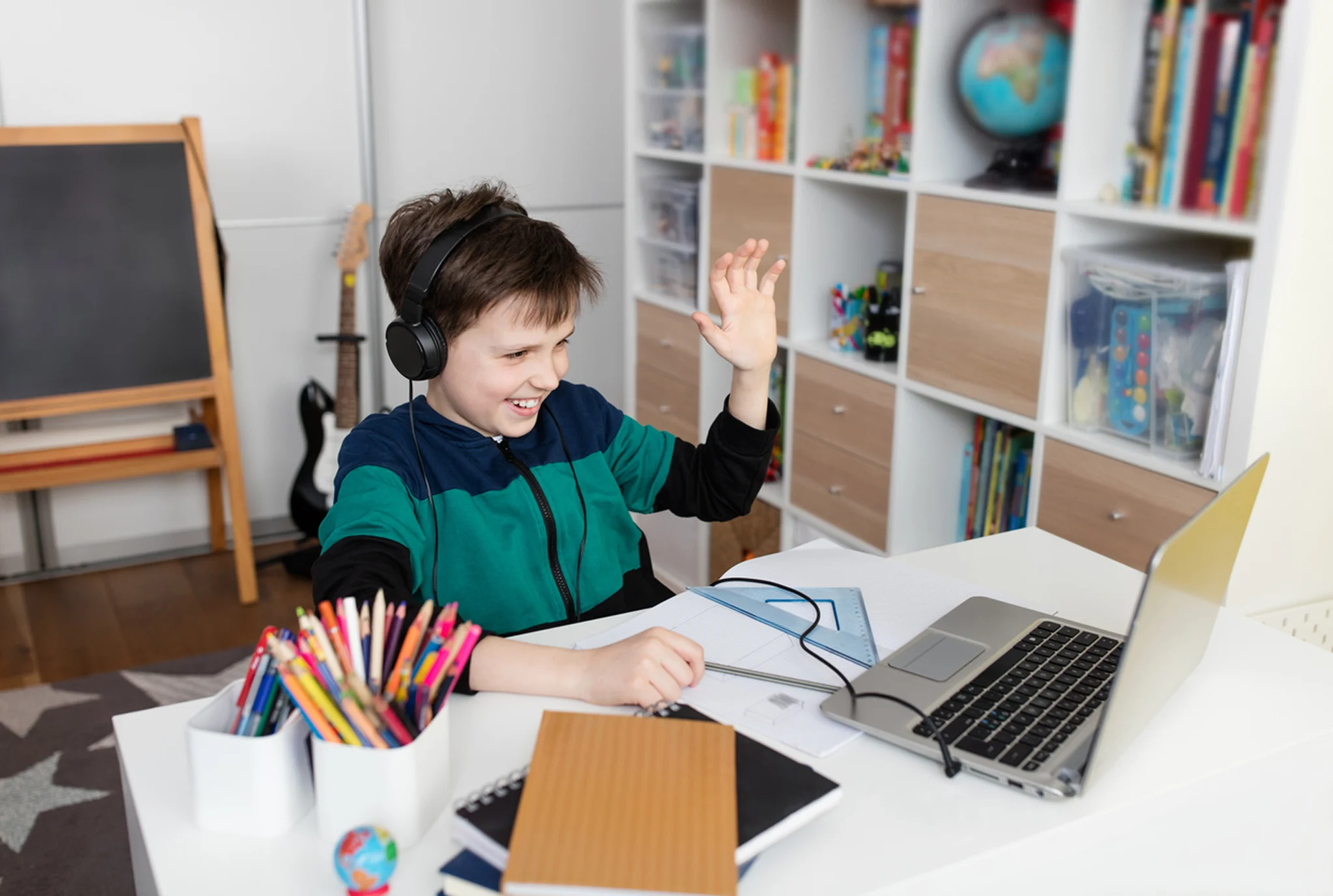 A young boy wearing headphones waves at his laptop during an online class. He's seated at a desk with colorful pencils, books, and a globe nearby, projecting a cheerful mood.