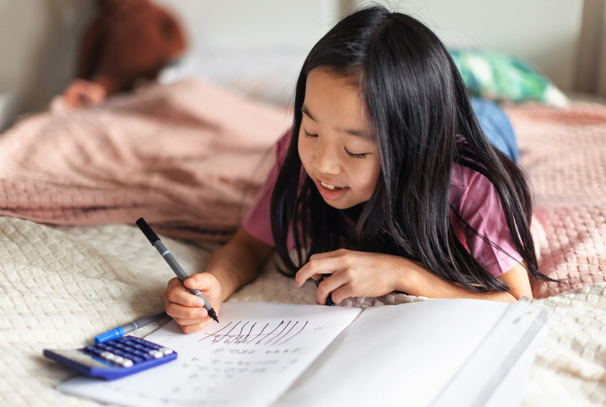 Niña acostada en la cama, haciendo la tarea con un bolígrafo, cuaderno y calculadora. Ella parece enfocada y contenta en un ambiente acogedor de dormitorio.
