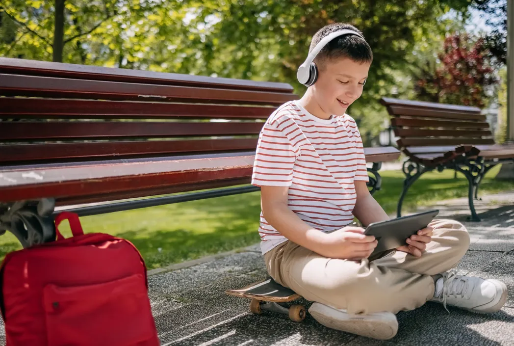 Un niño con auriculares y una camisa a rayas se sienta con las piernas cruzadas en una patineta, usando una tableta. Está en un parque junto a una mochila roja. Entorno relajado y soleado.