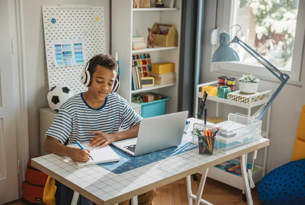 Un niño con auriculares estudia en un escritorio con una computadora portátil y una computadora portátil. La habitación es luminosa y organizada, transmitiendo un estado de ánimo concentrado y alegre.