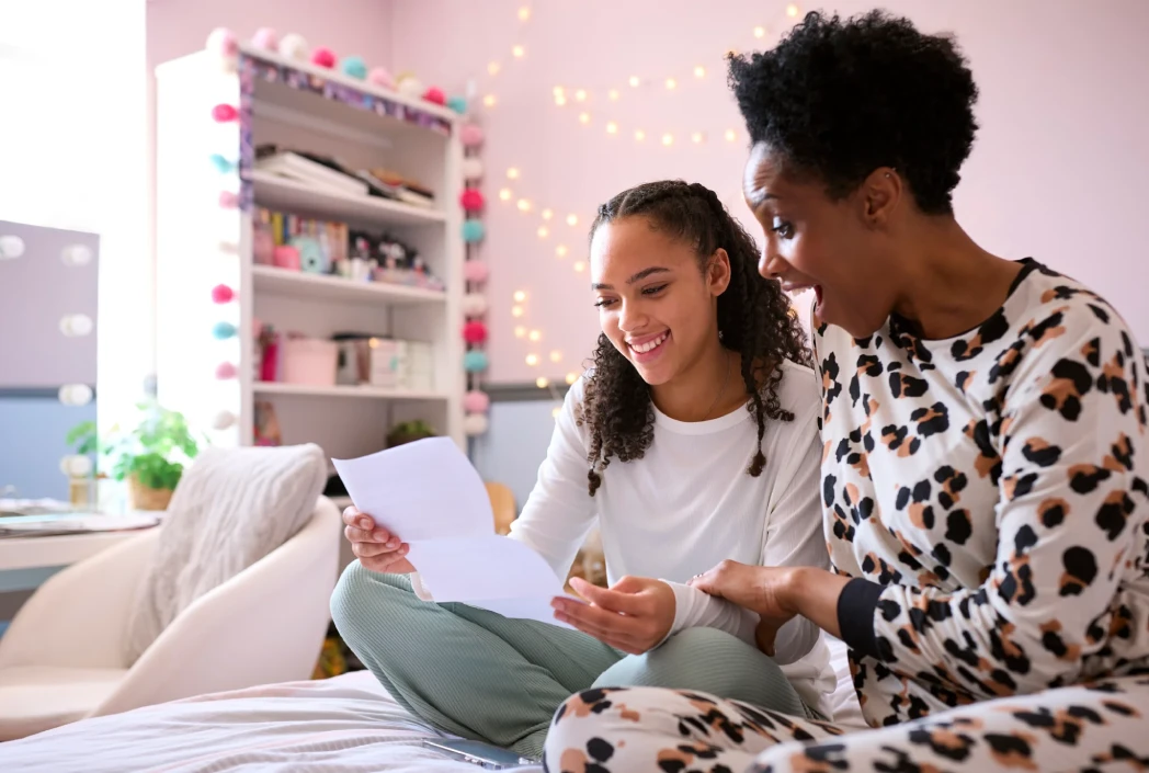 Una mujer y una niña se sientan en una cama en una habitación acogedora, leyendo alegremente una carta. La habitación está decorada con luces de hadas y coloridas guirnaldas de pompones.