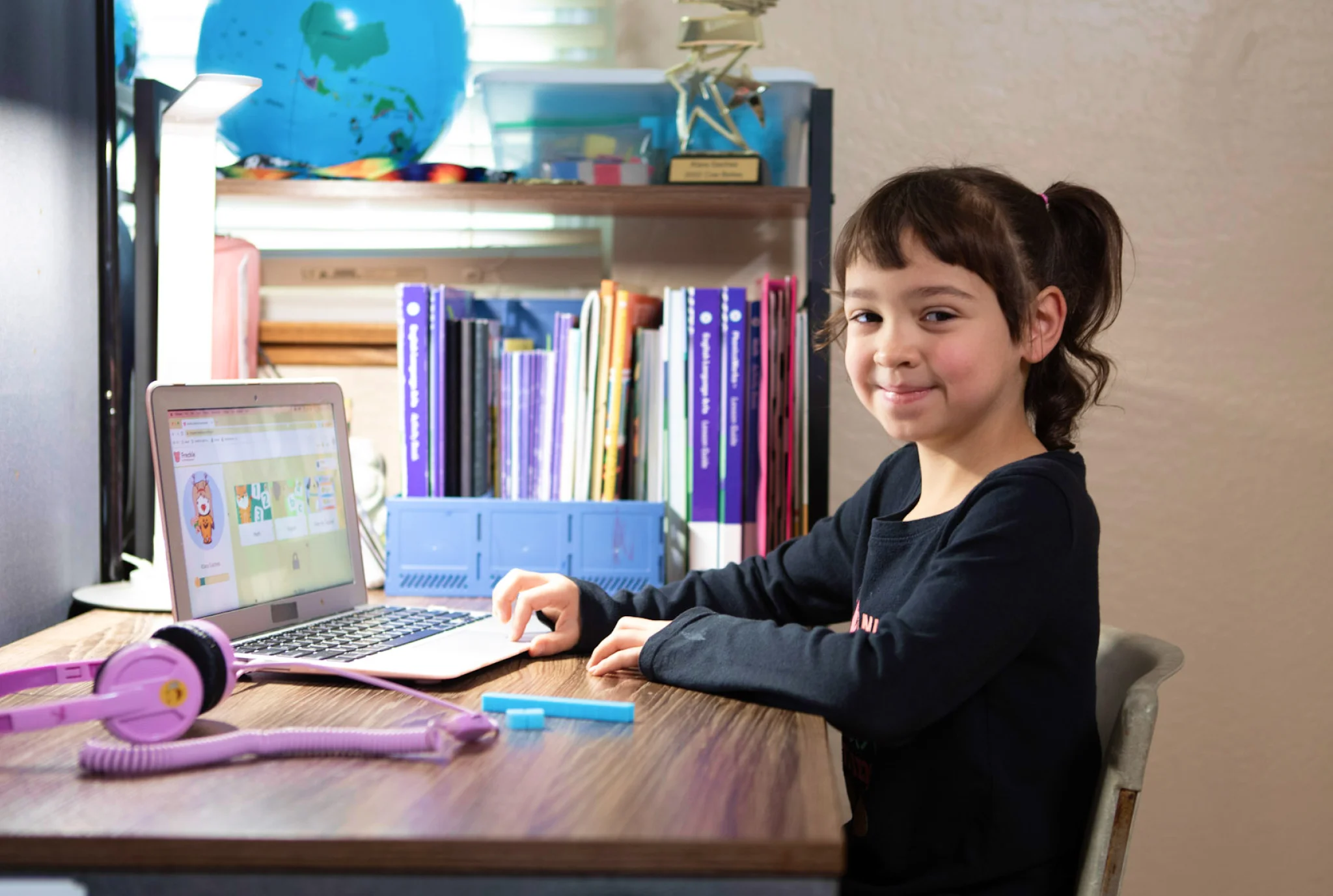 A young girl smiles while using a laptop at a wooden desk, surrounded by books and a globe. Purple headphones rest nearby, conveying a studious, cheerful mood.