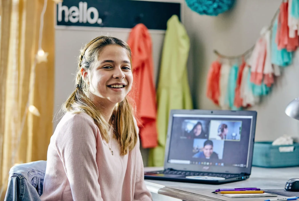 A smiling girl sits at a desk with a laptop open to a video call, in a colorful room decorated with hanging fabric. The atmosphere is cheerful and inviting.